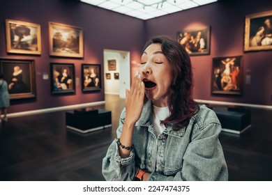a bored girl visitor or student in an art gallery or museum yawns and looks at the masterpieces of classical artists