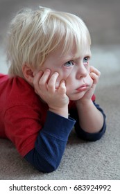 A Bored And Grumpy Toddler Child Is Laying On The Carpet At Home, Pulling On His Cheeks And Making A Pouting Face.