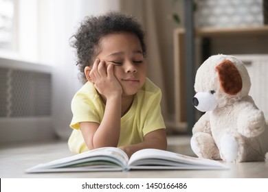 Bored Little Biracial Boy Lie On Warm Floor At Home Reading Book Together With Stuffed Teddy Toy, Funny Black Preschooler Child Distracted From Studying Dream Playing With Plush Friend In Living Room