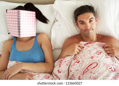 Bored Looking Man Lying In Bed Next To Woman Reading Book