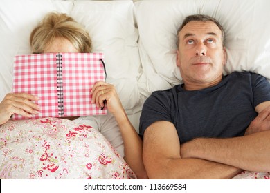 Bored Looking Senior Man Lying In Bed Next To Senior Woman Reading Book