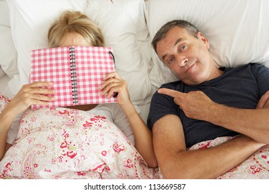 Bored Looking Senior Man Lying In Bed Next To Senior Woman Reading Book