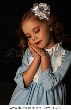 Bored lovely little doll cover girl in blue dress at black background, pensive closed eyes. Darling child of image isolated with light and shadow, studio shot. Fashion style kid concept. Copy ad space