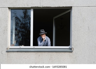 Bored Man In Corona Quarantine Looking Out Of Window To The Street.Man With The Hat Celebrating Alone Birthday At Home During Covid-19 Lockdown. Sad Birthday During Corona Quarantine. 