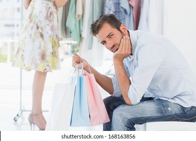 Bored man sitting with shopping bags while woman by clothes rack in the background - Powered by Shutterstock