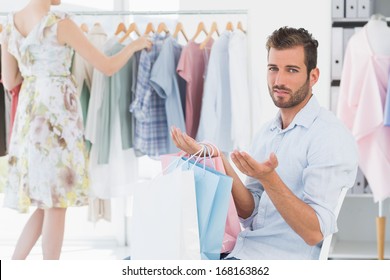 Bored Man Sitting With Shopping Bags While Woman By Clothes Rack In The Background