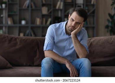 Bored millennial generation man sitting on sofa, feeling lonely at home. Unhappy young male suffering from negative thoughts, having depressive mood or problems in relations, regretting mistakes.