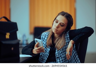 
Bored Office Worker Checking her Phone Sitting at Desk. Lazy procrastinator employee being distracted by doom scrolling

