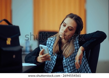 
Bored Office Worker Checking her Phone Sitting at Desk. Lazy procrastinator employee being distracted by doom scrolling
