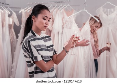 So Bored. Sad Cheerless Woman Waiting For Her Friend While Drinking Sparkling Wine In The Wedding Dress Salon