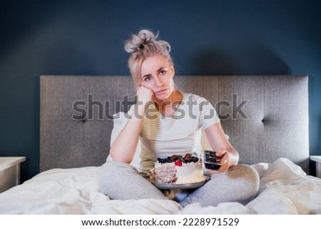 Bored, sad woman in home clothes eating cake and using the remote control to change the channel while watching television in bed at night. Overeating, Jamming of negative emotions. Selective focus.
