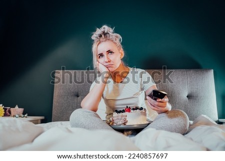 Bored, sad woman in home clothes eating cake and using the remote control to change the channel while watching television in bed at night. Overeating, Jamming of negative emotions. Selective focus