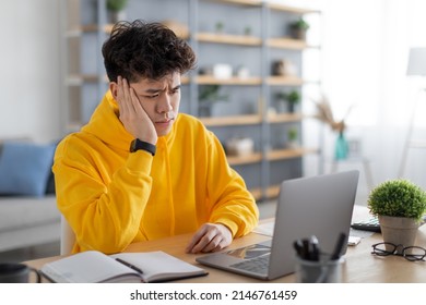 Bored Sad Young Asian Guy Sitting At Desk Using Laptop, Looking At Pc Screen, Thinking About Problems During Online Lesson Or Remote Work At Home Office. Tired Man Leaning Head On Hand, Watching News