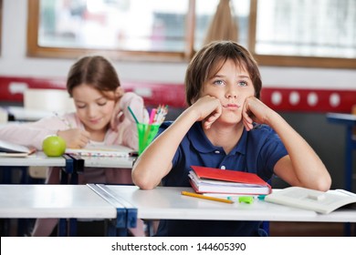 Bored schoolboy looking away while sitting at desk with girl in background at classroom - Powered by Shutterstock