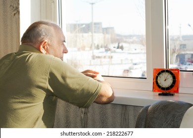 Bored Senior Man Waiting Patiently For Time To Pass As He Stands Staring Disconsolately Out Of A Window On A Sunny Day Waiting For Someone To Arrive For A Visit