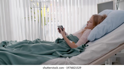 Bored small girl in bed in hospital using tablet to pass time. Side view of sick kid patient with nasal tube and catheter watching cartoon on digital tablet lying in hospital bed
