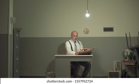 Bored Vintage Style Businessman Sitting At A Small Desk, Reading A Newspaper In A Cluttered Messy Office With Cheap Furniture