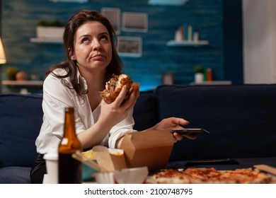 Bored Woman Browsing Social Media On Smartphone Eating Delicious Burger Takeout Dinner Sitting On Sofa In Home Living Room. Caucasian Female Having Takeaway Dinner Junk-food Order.