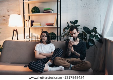 bored woman sitting on couch with arms crossed while man using smartphone in living room