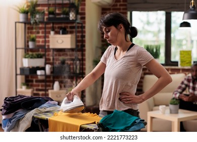 Bored woman tired of ironing clothes at home watching in frustration while doing household chores. African American husband in background resting while wife does all the work. - Shutterstock ID 2221773223