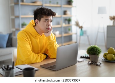 Un chico asiático aburrido sentado en su escritorio usando una laptop pc, mirando a un lado, pensando en algo durante una lección en línea o en un trabajo remoto en su oficina. Hombre cansado con un trabajo aburrido y distante, que se siente perezoso