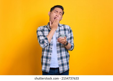 Bored Young Asian Man In Plaid Shirt Holding Mobile Phone And Feeling Lack Of Sleep Isolated On Yellow Background