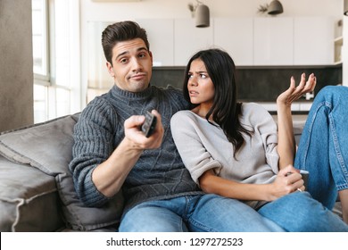 Bored Young Couple Sitting On A Couch At Home, Watching TV, Drinking Tea