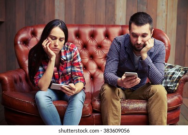 Bored Young Couple Sitting On Couch And Looking At Their Phones