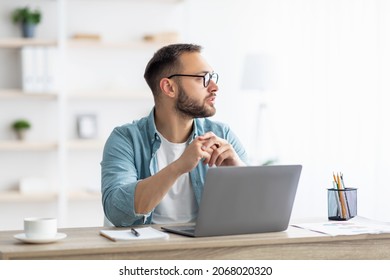 Bored Young Guy Sitting Near Laptop, Looking Aside, Daydreaming, Thinking About Something During Online Work At Home Office. Millennial Caucasian Man Having Dull Remote Job