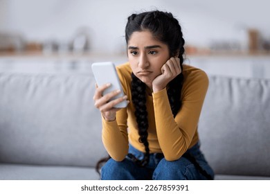 Bored Young Indian Woman Looking At Smartphone Screen While Sitting On Couch At Home, Disappointed Millennial Hindu Female Holding Mobile Phone, Waiting For Important Call Or Sms, Copy Space
