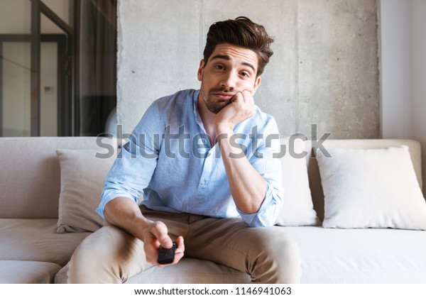 Bored young man holding tv remote control while\
sitting on a couch at\
home