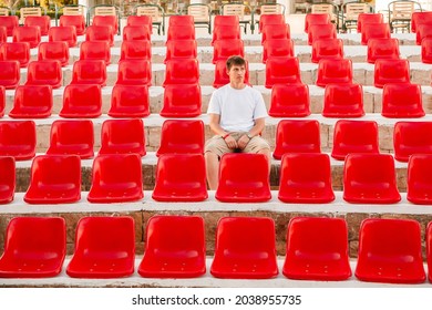 Bored Young Man Sitting In Deserted Audience With Bright Red Seats. Absence Of People In Open-air Amphitheater Due To Coronavirus COVID-19 Pandemic Lockdown And Quarantine.