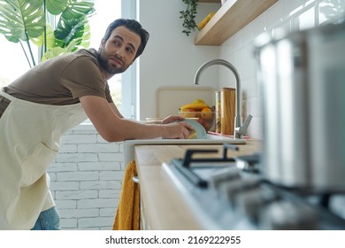 Bored young man washing dishes at the domestic kitchen and looking over shoulder - Powered by Shutterstock