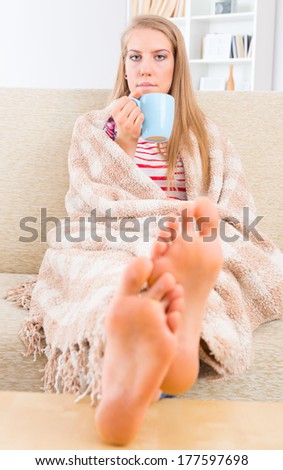 Bored young woman covered with blanket sitting on the sofa at home with feets on the table