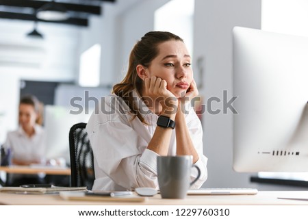 Bored young woman dressed in shirt sitting at her workplace at the office, looking at computer