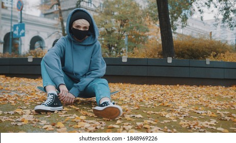 Bored young woman with face mask and hoodie sitting on the skateboard in the city park in autumn. Social distance during coronavirus outbreak. High quality photo - Powered by Shutterstock