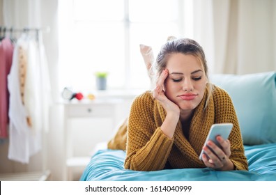 Bored Young Woman With Smartphone Lying On Bed Indoors At Home.