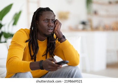 Boring Program. Portrait Of Bored Young Black Guy Watching TV At Home, Discontented African American Man Sitting On Couch At Home, Leaning Head On Hand And Switching Channels With Remote Controller