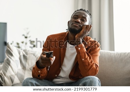 Boring television program. Bored black man watching TV and switching channels with remote control, sitting on couch. Discontented male viewer watching uninteresting movie