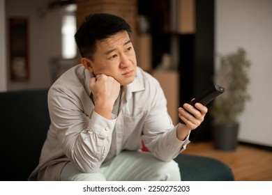 Boring television program concept. Bored mature japanese man watching TV, switching channels with remote control, sitting on sofa at home. Discontented male viewer watching uninteresting movie