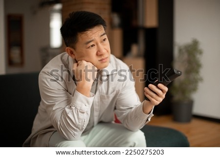 Boring television program concept. Bored mature japanese man watching TV, switching channels with remote control, sitting on sofa at home. Discontented male viewer watching uninteresting movie