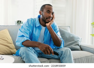 Boring Television Program. Discontented Young Black Guy Watching TV, Switching Channels With Remote Control, Sitting On Sofa At Home And Feeling Lonely, Free Copy Space