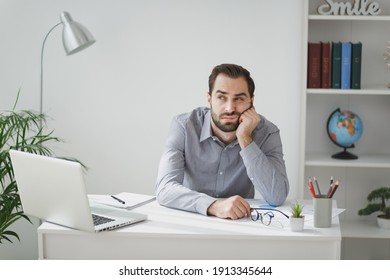 Boring young bearded business man in gray shirt sitting at desk working on laptop pc computer in light office on white wall background. Achievement business career concept. Put hand prop up on cheek