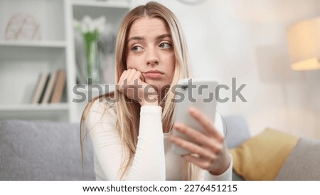 Boring young blond woman scrolling feed of social networks products in internet store on smartphone in the living room Tired exhausted female look at mobile screen without enthusiasm at home Сток-фото © 