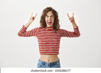 Born To Rock This World. Joyful Woman In Fashionable Clothes Screaming Out Loud And Showing With Raised Arms Horns Or Rock Gesture, Closing Eyes, Expressing Excitement Of Being On Concert Of Band