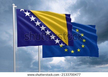 BosniaHertzegovina and EU flag together next to each other on a flagpole. Bosnia and Hertzegovina flag in front of a European Union flag on a dramatic stormy sky background