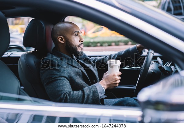 Bossy confident African American\
executive man in formal clothes with takeaway hot drink focusing\
and driving automobile standing in traffic jam in\
downtown