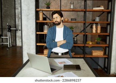 Bossy successful financial expert or trader standing next to working table with laptop and paper documents with crossed hands in loft style home interior, satisfied with his new achievements