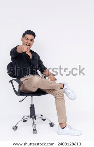 A bossy young man pointing to the camera while berating at someone. Looking irate and angry. Sitting cross legged on an office chair. Studio shot isolated on a white background.