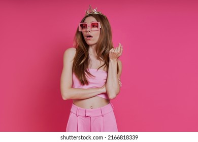 Bossy young woman in funky crown looking displeased while standing against pink background
